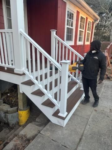 Person building stairs of house with white railing and brown steps. Red house in the background.