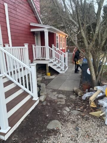 Red house with white porch and stairs, leading to a concrete path. Person standing near a tree.