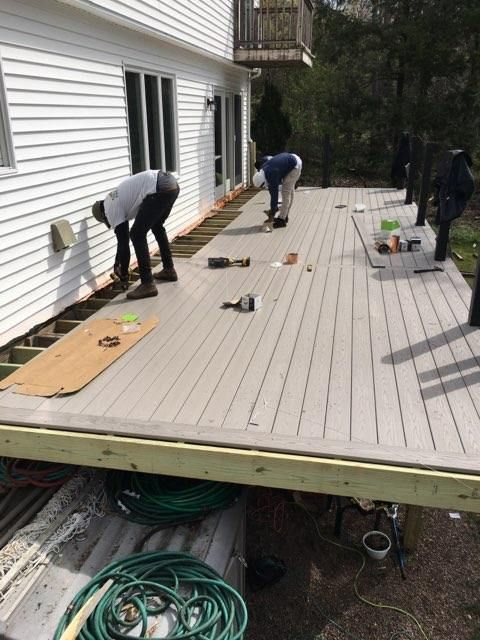 Workers installing gray composite deck boards on a wood frame deck next to a white house.