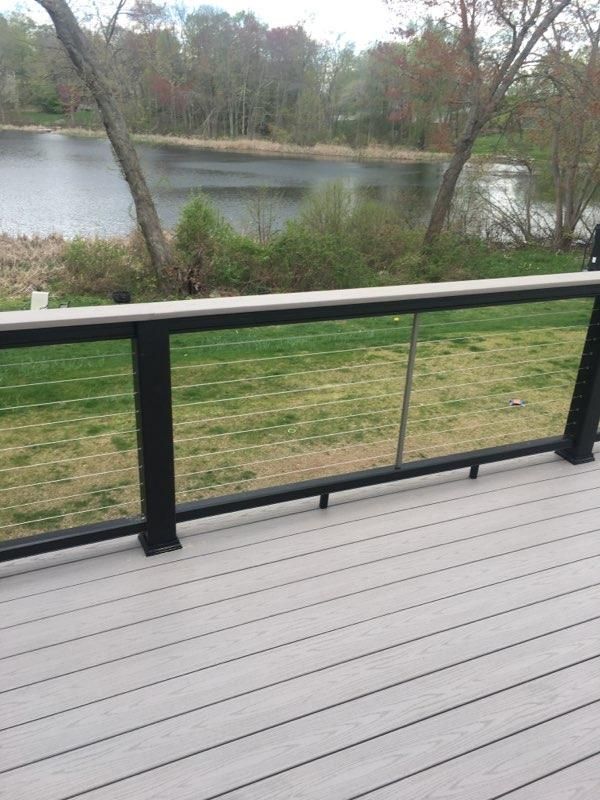 Deck with glass railing overlooking a lake. Green grass, trees, and cloudy sky in the background.