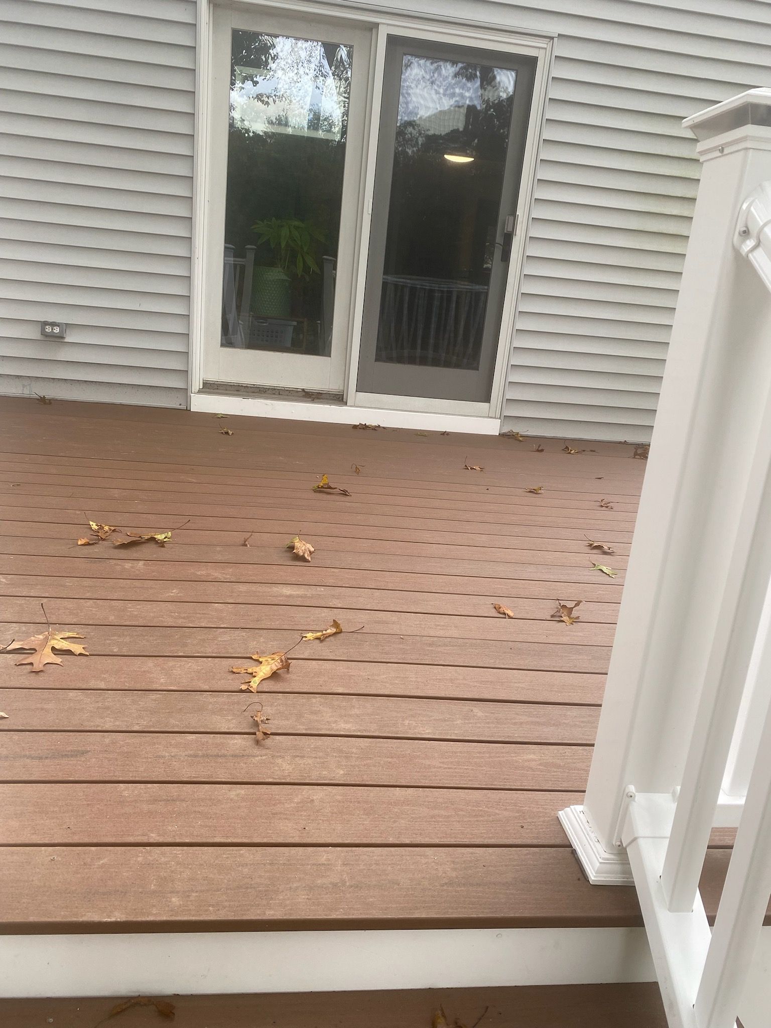 Brown deck with scattered leaves, white railing, and glass doors to a house.