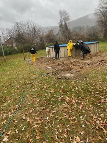 Four people in rain gear stand near a partially dismantled above-ground pool in a grassy yard under a gray sky.
