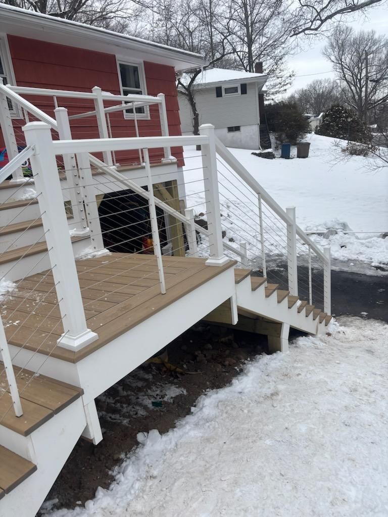 White deck with cable railings and stairs in a snowy setting next to a red house.