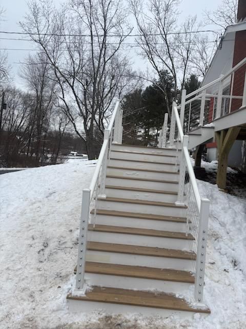 Outdoor staircase with white railings and wooden steps covered in patches of snow, leading up to a red building.