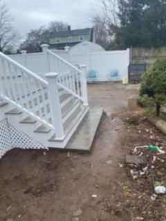 White deck with stairs, leading to a muddy yard with a white fence and a shed.