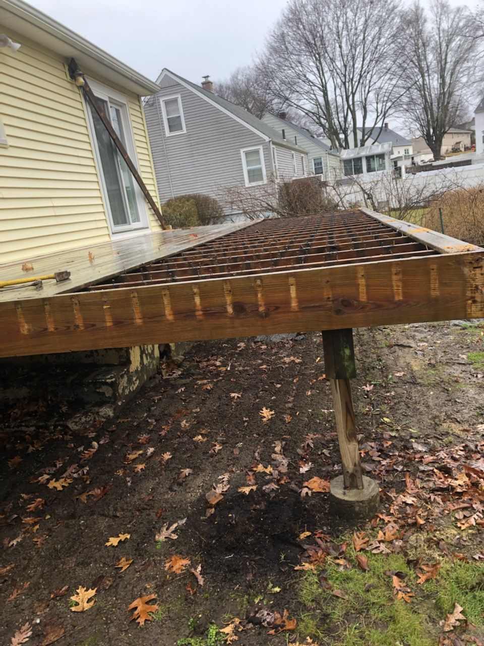 Damaged wooden deck attached to a yellow house, supported by posts. Overcast day.