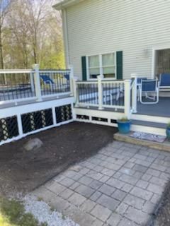Deck with white railing, black lattice, and gray pavers. Next to a house with green shutters and blue chairs.