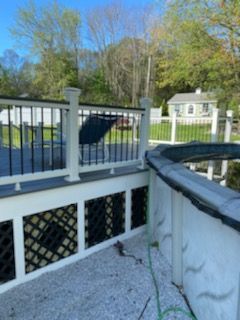 Deck with black railings, lattice skirting, and an above-ground pool. Gravel ground and greenery in the background.