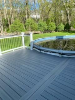 Gray deck surrounds a pool, with black railing and green trees in the background.
