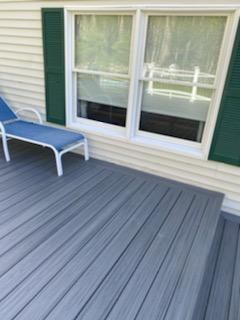 Blue deck with a lounge chair next to a wall with white framed windows and green shutters.