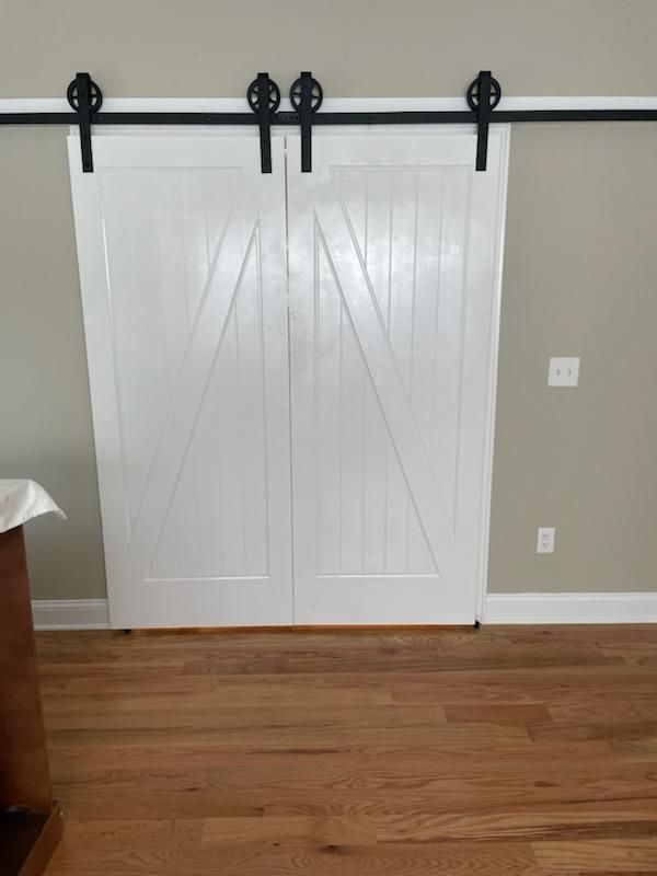 Two white barn doors on black track, wooden floor, gray wall.