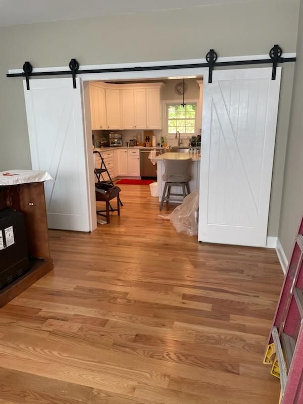 White barn doors on a black track, opening to a kitchen with white cabinets and a window, hardwood floor.
