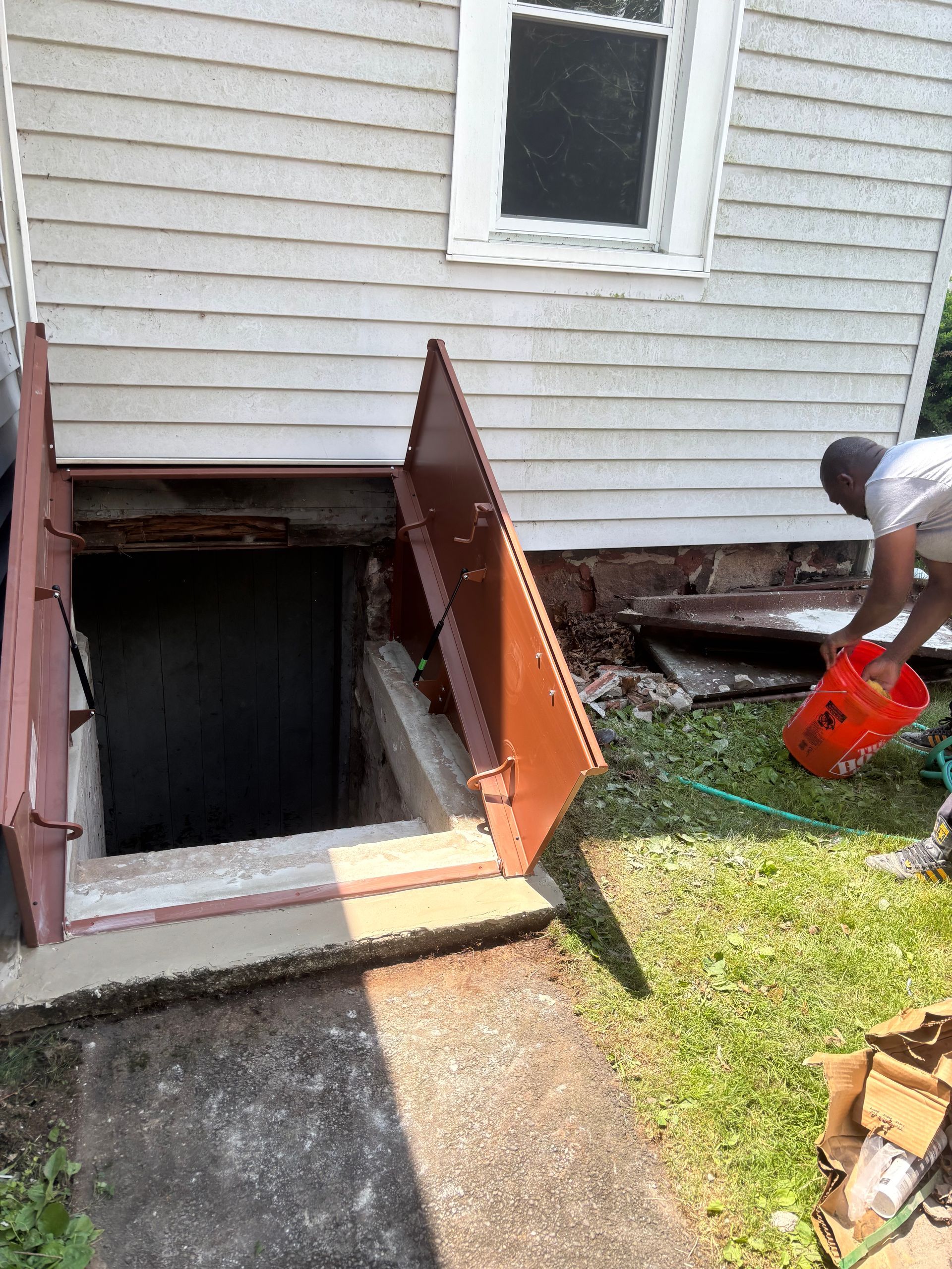 Basement entrance with open brown door. Man pours water near the opening, concrete steps, house siding.