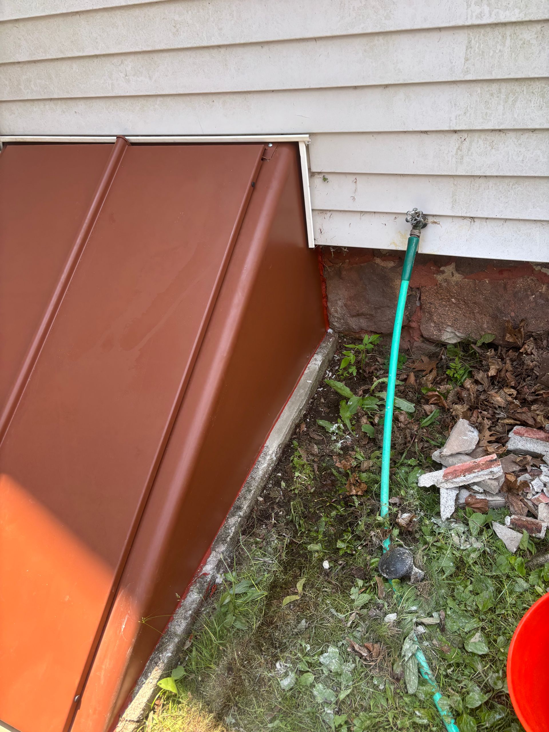 Brown metal cellar door with a green hose and grass. The side of a white house is visible.