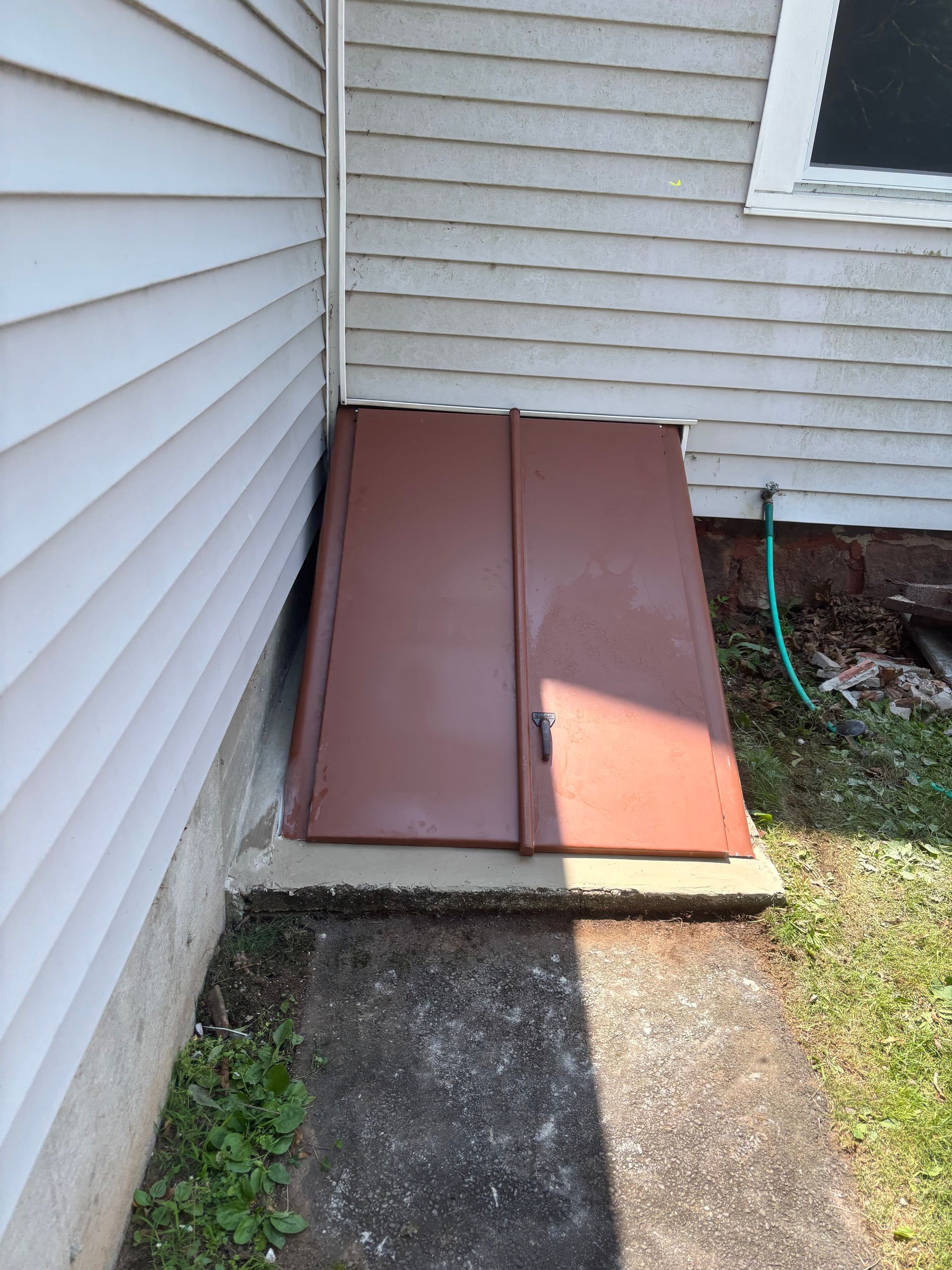 Brown cellar doors next to a white house with a concrete slab and a green hose nearby.