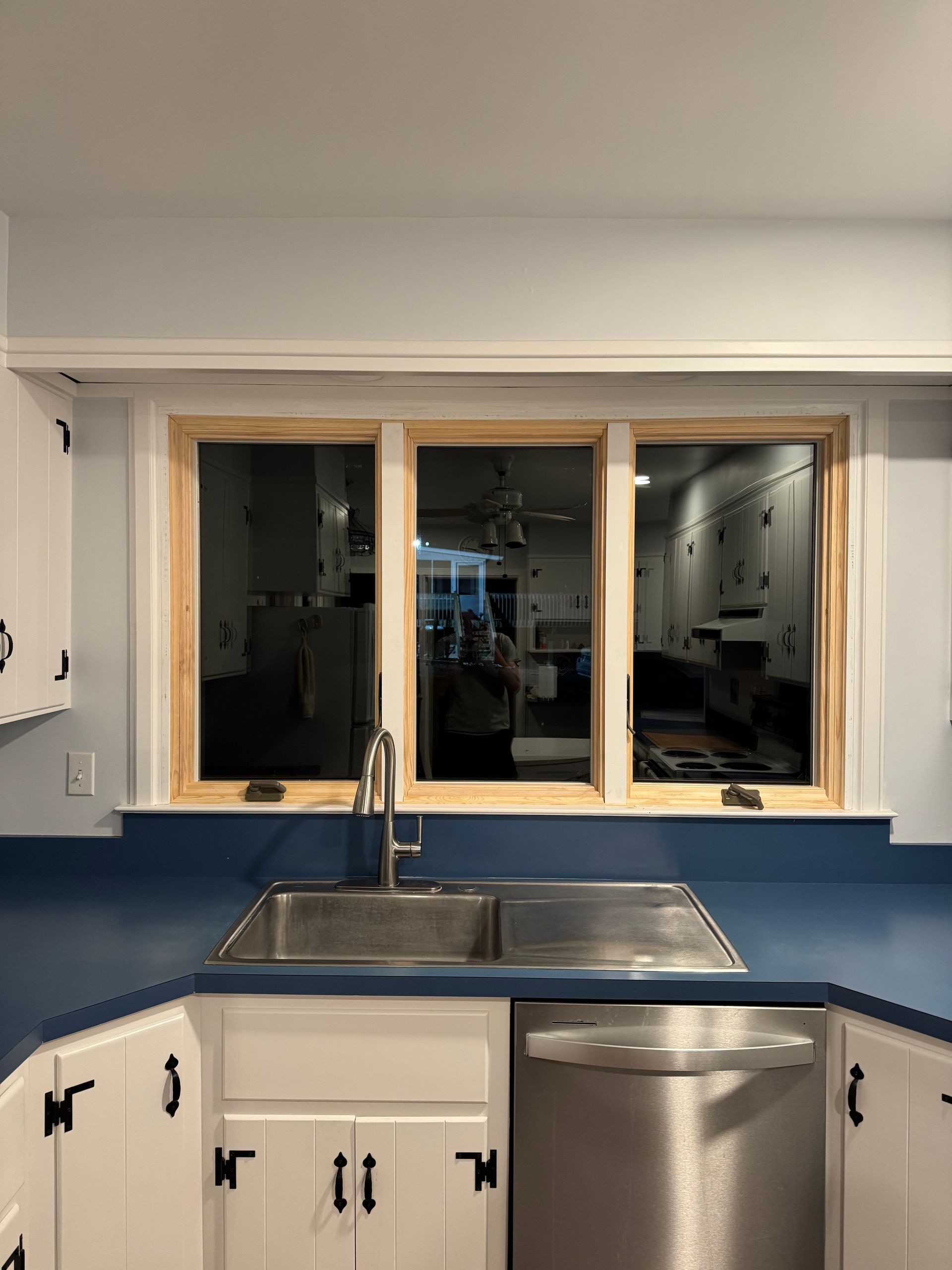 Kitchen with triple window above a stainless steel sink and blue countertop. White cabinets with black hardware.