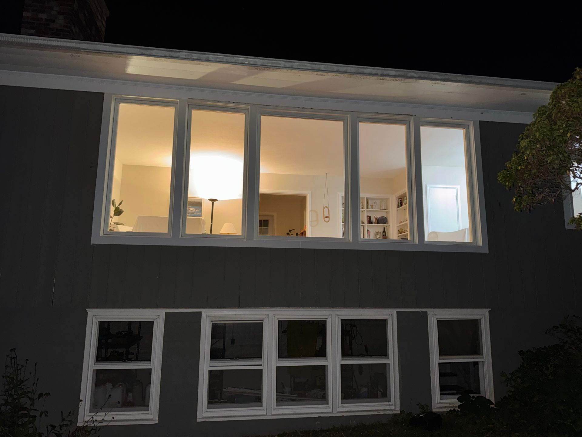 A dark house exterior at night with lit windows, showing a living space inside.