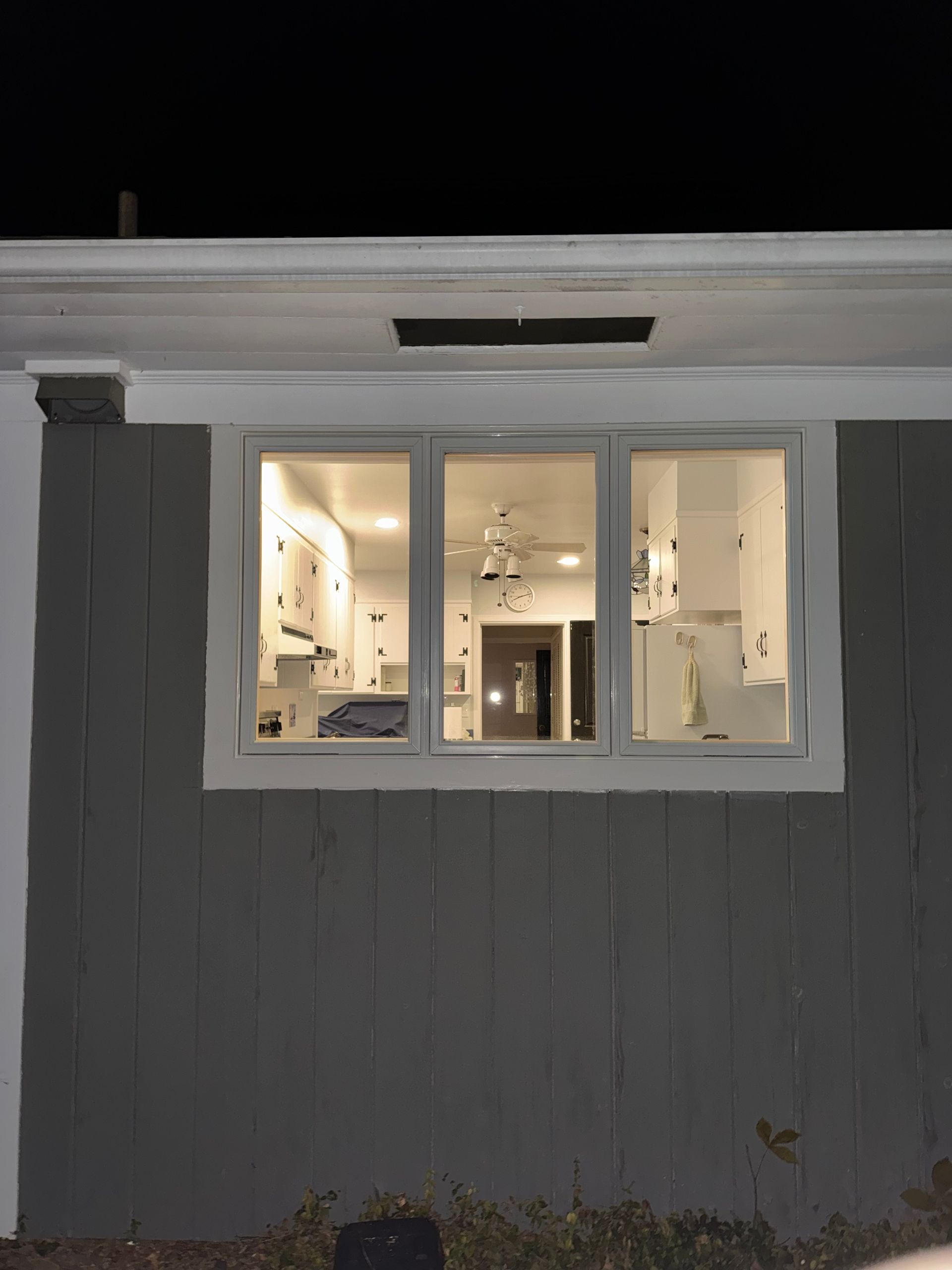 Exterior view of a house at night, illuminated interior visible through three windows, showing a kitchen.