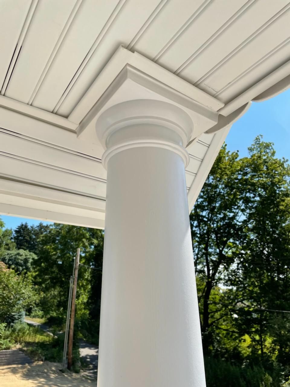 White painted porch column and ceiling details against a bright sky, with trees visible in the background.