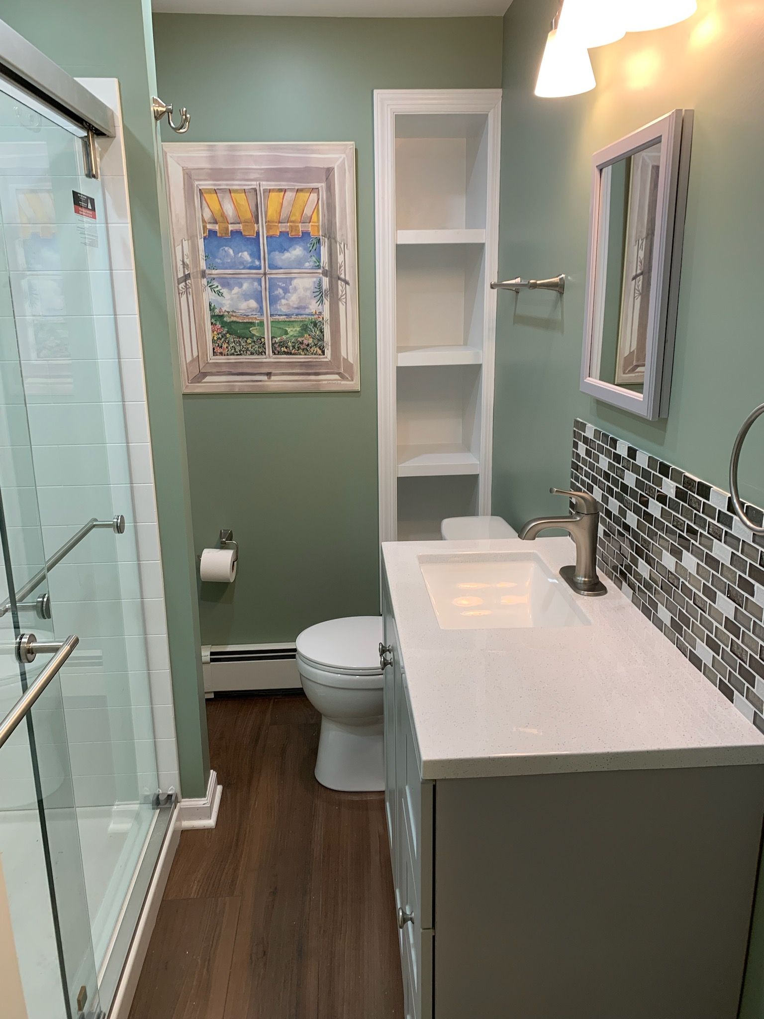 Bathroom with a shower, toilet, sink, and shelves. Sage green walls, wood-look floor, and a framed window art piece.