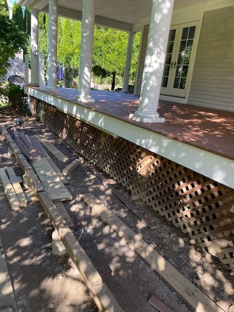 Porch with lattice skirting; construction materials on ground. White columns, brown decking, sunny outdoor setting.