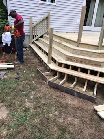 Man builds wooden deck stairs next to a house. Partially constructed stairs, tools, and materials are visible.