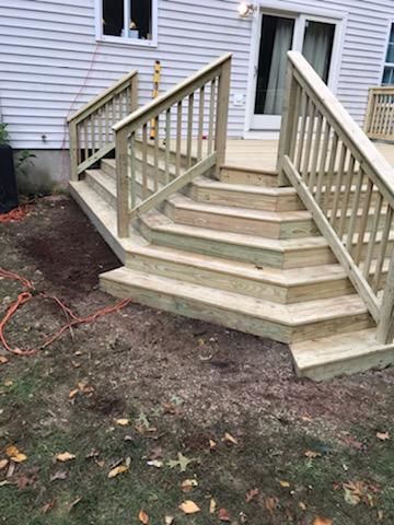 Wooden deck with stairs leading to a house, surrounded by grass and dirt.