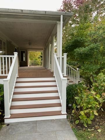A covered porch with white columns, stairs with brown treads, and a concrete path. Lush greenery surrounds it.