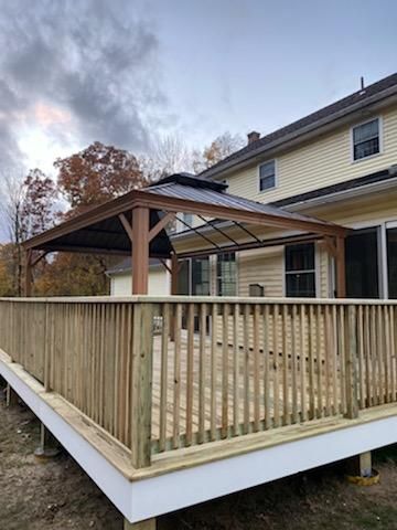 Wooden deck with gazebo attached to a yellow house with a cloudy sky backdrop.