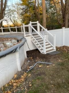 White staircase leading down from a deck beside a pool, surrounded by a white fence and trees.