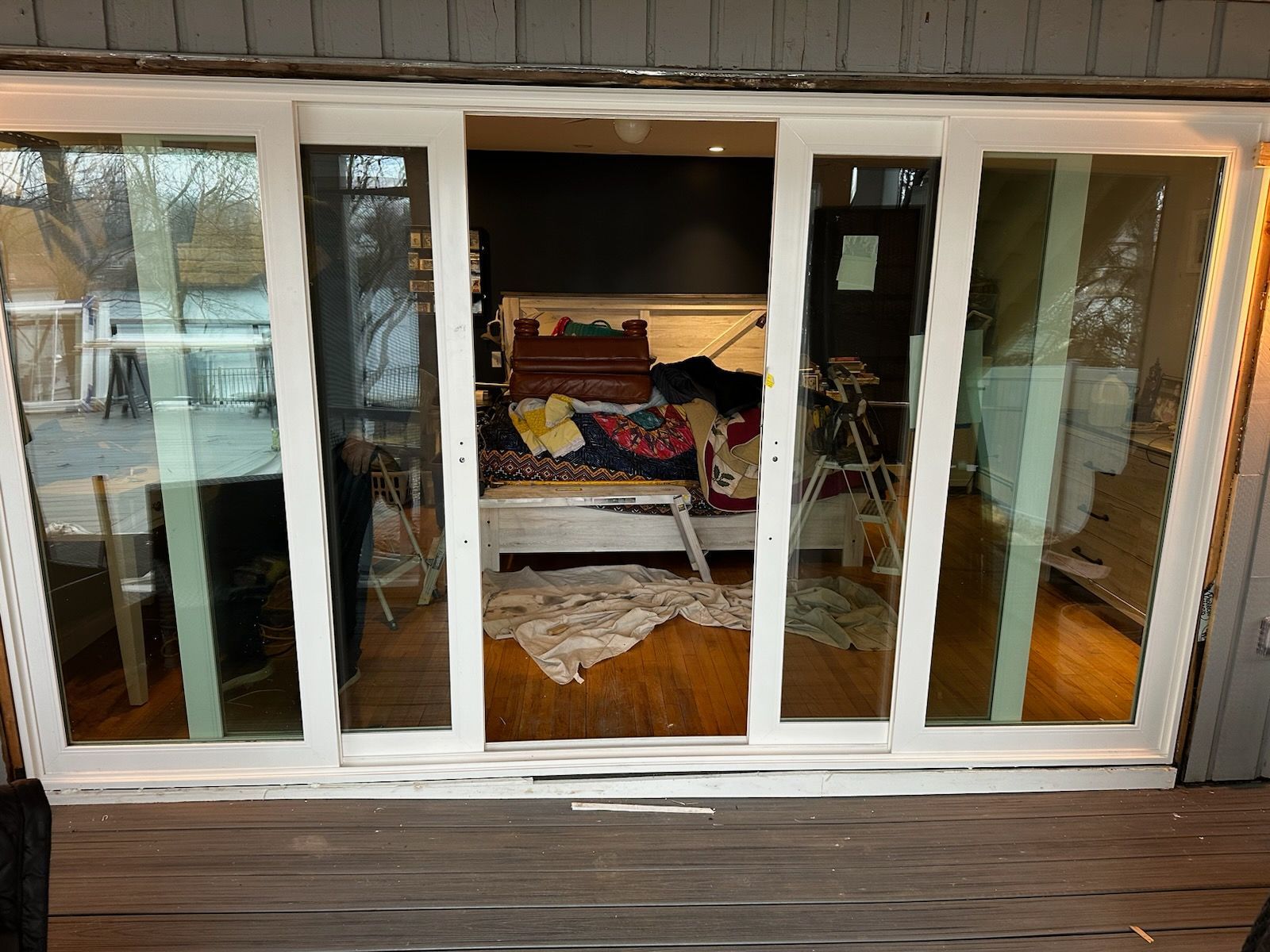 Sliding glass doors open to a messy room with a bed. The doors are framed in white, on a wooden deck.