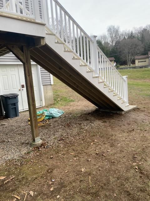 Wooden deck stairs leading to ground, white railing, brown dirt ground, garage in background.