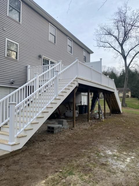 A two-story house with a white deck and staircase. Wooden support beams stand below the deck. Backyard setting.