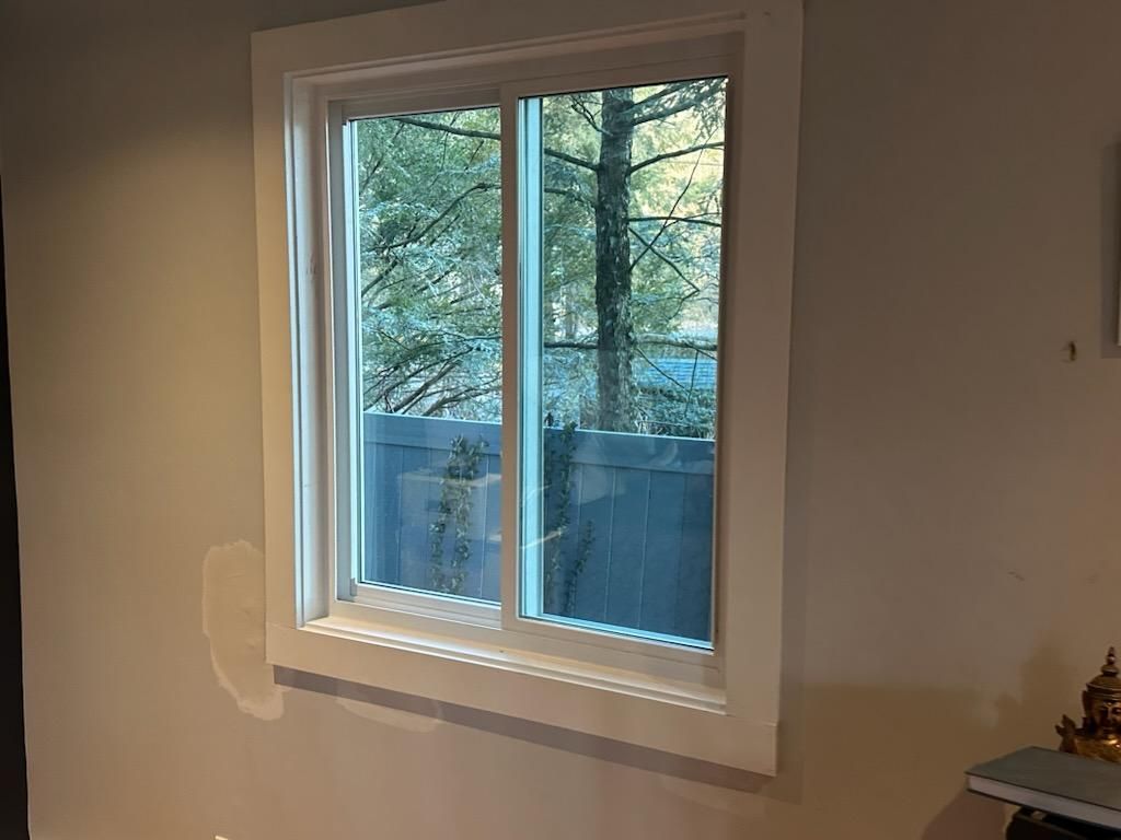 A white-framed window on a beige wall, looking out onto a wooded area and a fence.