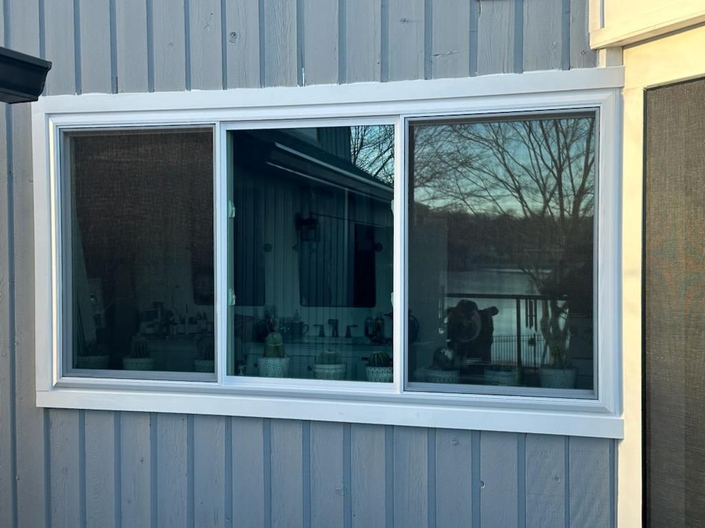 White-framed triple-pane window set in light-blue siding; reflects outdoor scene, including trees and a building.