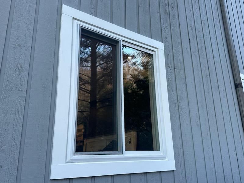 White-framed sliding window on gray wooden siding, reflecting trees.