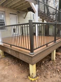 Brown composite deck with black metal railing, supported by yellow posts on a dirt surface.