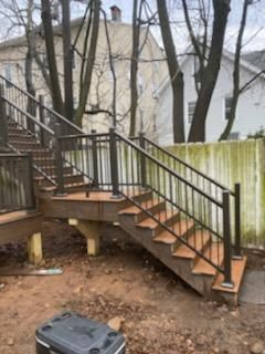 Outdoor wooden staircase with black railing leads to a deck. Surrounding is bare ground, trees, and a weathered fence.