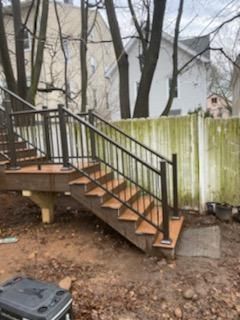 Wooden staircase with black railings, leading up to a deck, against a weathered fence and trees.