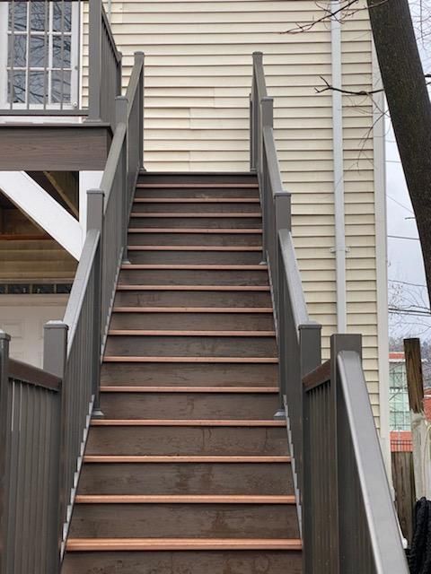 Outdoor wooden staircase leading upwards. Gray railings, brown steps. House exterior in background.