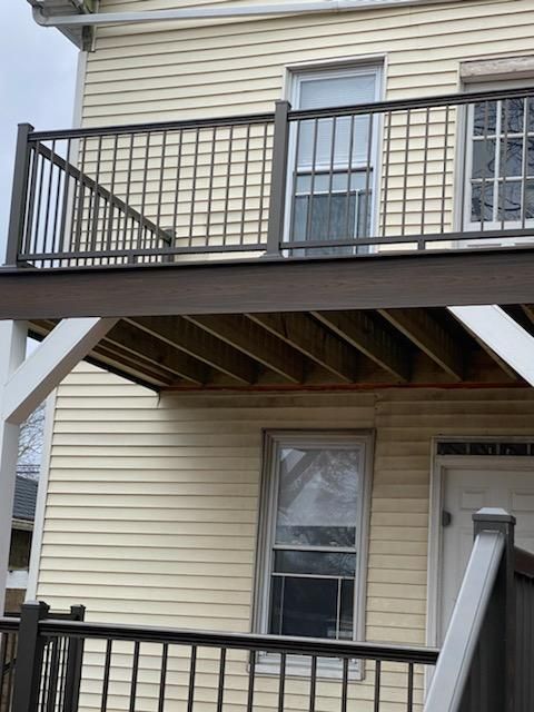 Two-story deck with dark railing and brown planks, attached to a yellow-sided house with windows and a door.