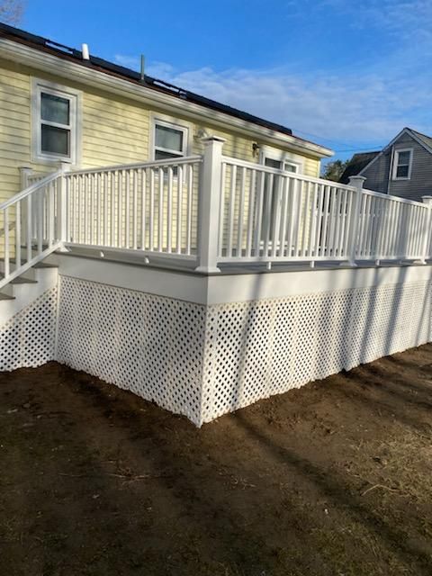 White deck attached to a yellow house with lattice skirting and railing, on a sunny day.