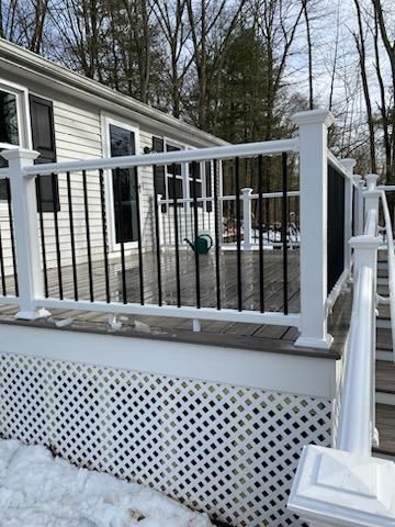 White deck with black railing and lattice. A watering can sits on the deck, surrounded by snow and trees.
