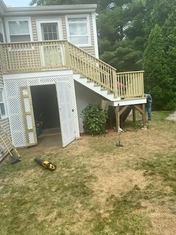 Wooden deck with stairs leading to a second-story landing. White lattice structure underneath with open doors, person working.