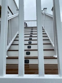 White stairs with brown treads, lit by square lights, ascending from a wooden deck. Railings flank the steps.