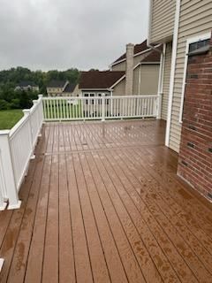 Wooden deck with white railing, next to a beige house with a brick accent, overlooking green grass and trees.
