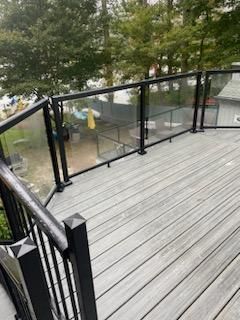 Gray wooden deck with black railing and glass panels, overlooking a body of water.