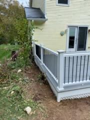 White deck with black vertical railings, beside a yellow house. Green vegetation surrounds.