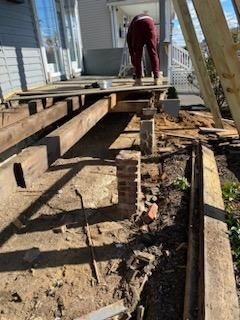 Construction of a deck. A person works on wooden beams. The ground is dirt and brick.