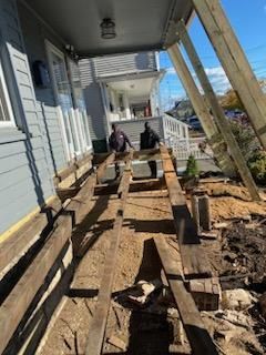 Workers renovating a porch with removed wooden planks. Supports hold up the porch roof. Soil is exposed.
